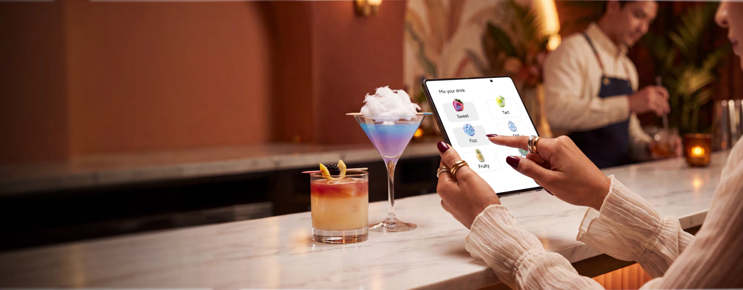 Client concept art: a guest orders on a Galaxy foldable at a marble bar, a layered martini and a sunset cocktail beside her and a bartender behind.